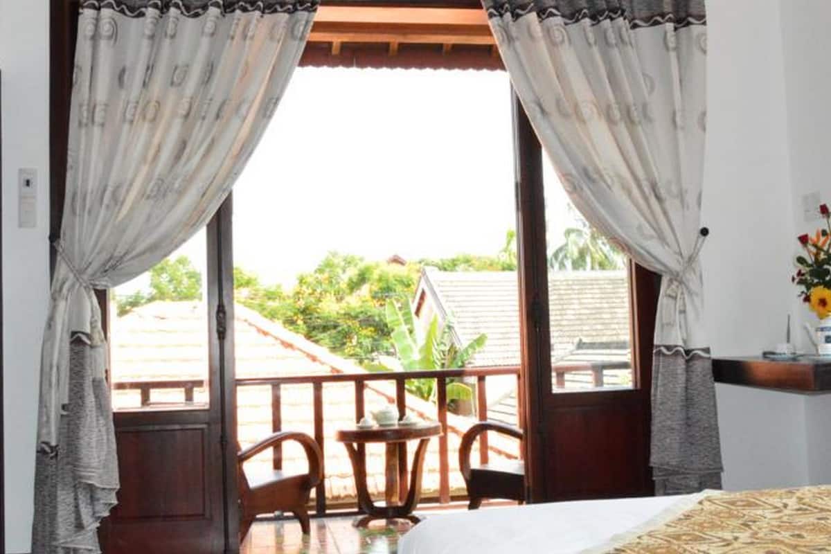 A room with a bed, a small table, and chairs. There is a balcony with a railing and a view of greenery outside.
