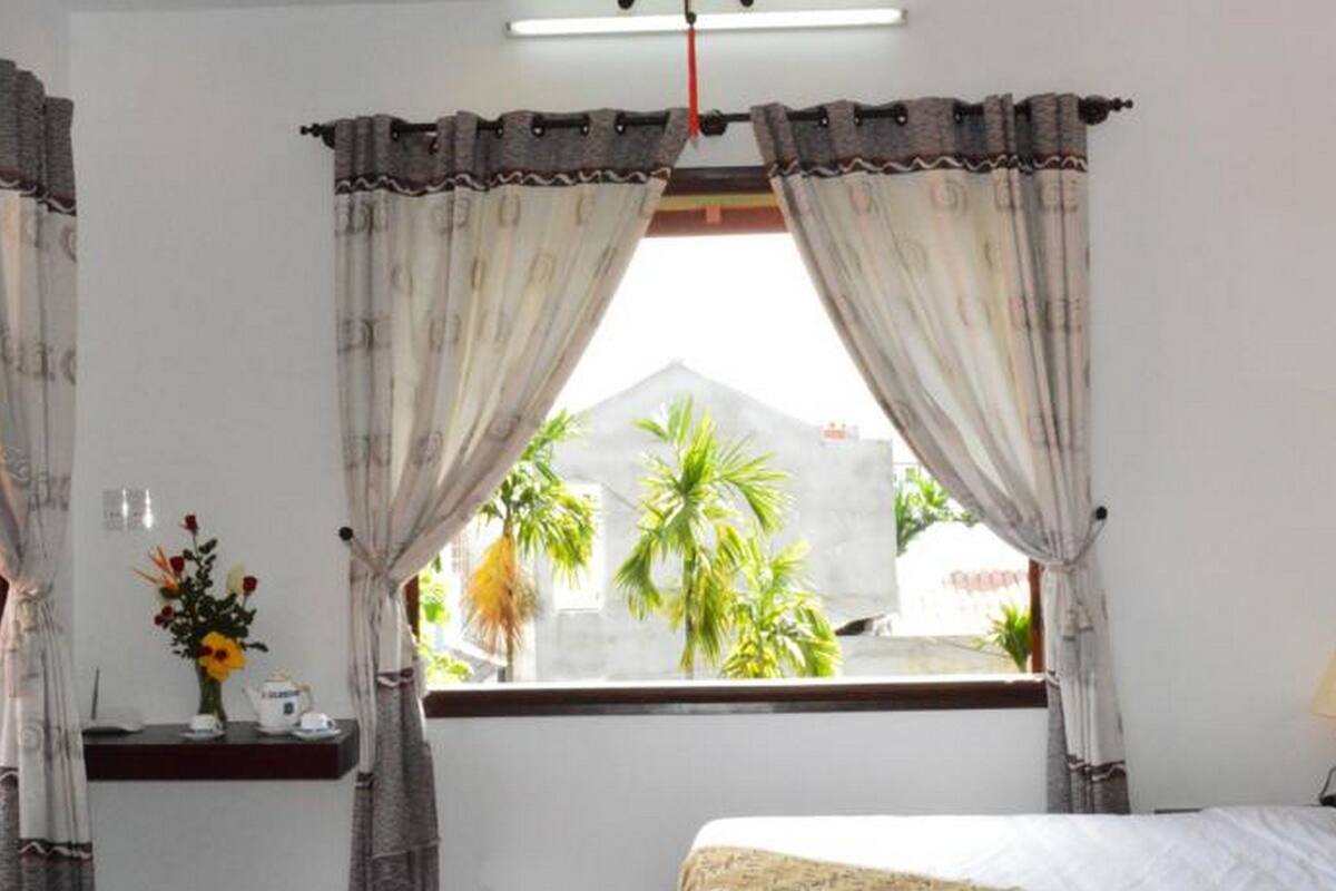 A bedroom with a bed, a window with curtains, a small table with a vase of flowers, and a view of palm trees outside.