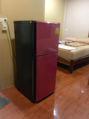 A compact room with a two-door refrigerator, a bed with a beige mattress and pillows, and a wooden floor.