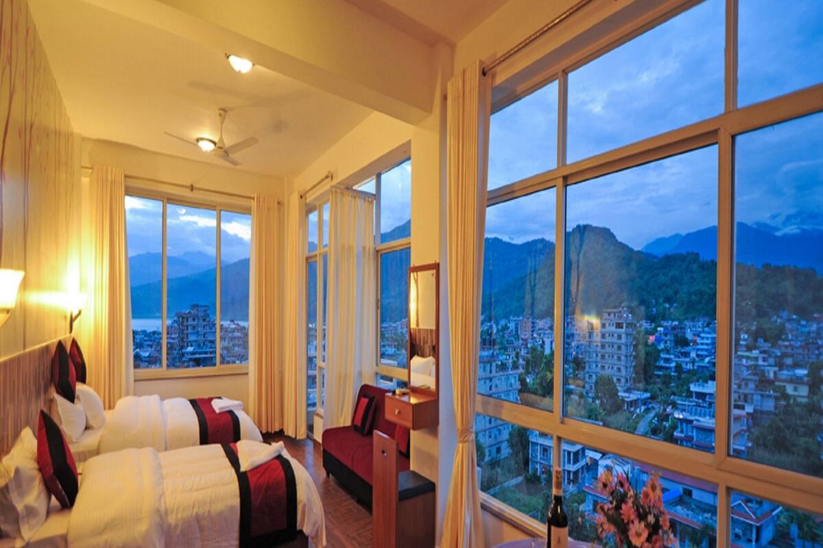 A hotel room with two beds, a desk, a round table with a bottle and glasses, and large windows offering a city view.