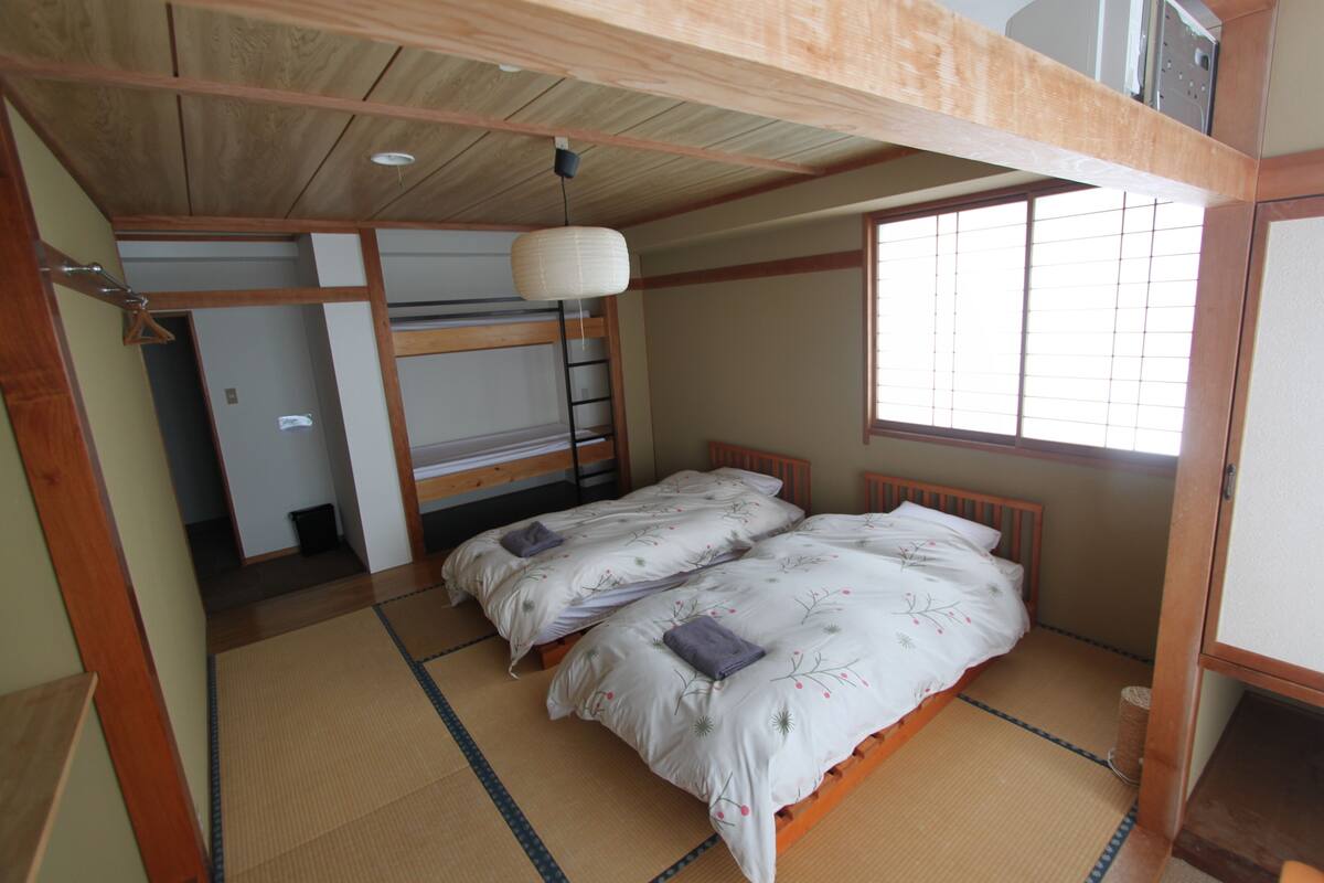A traditional Japanese-style room with tatami flooring, a low wooden bed with a futon, a hanging paper lantern, and a sliding door leading to another room.
