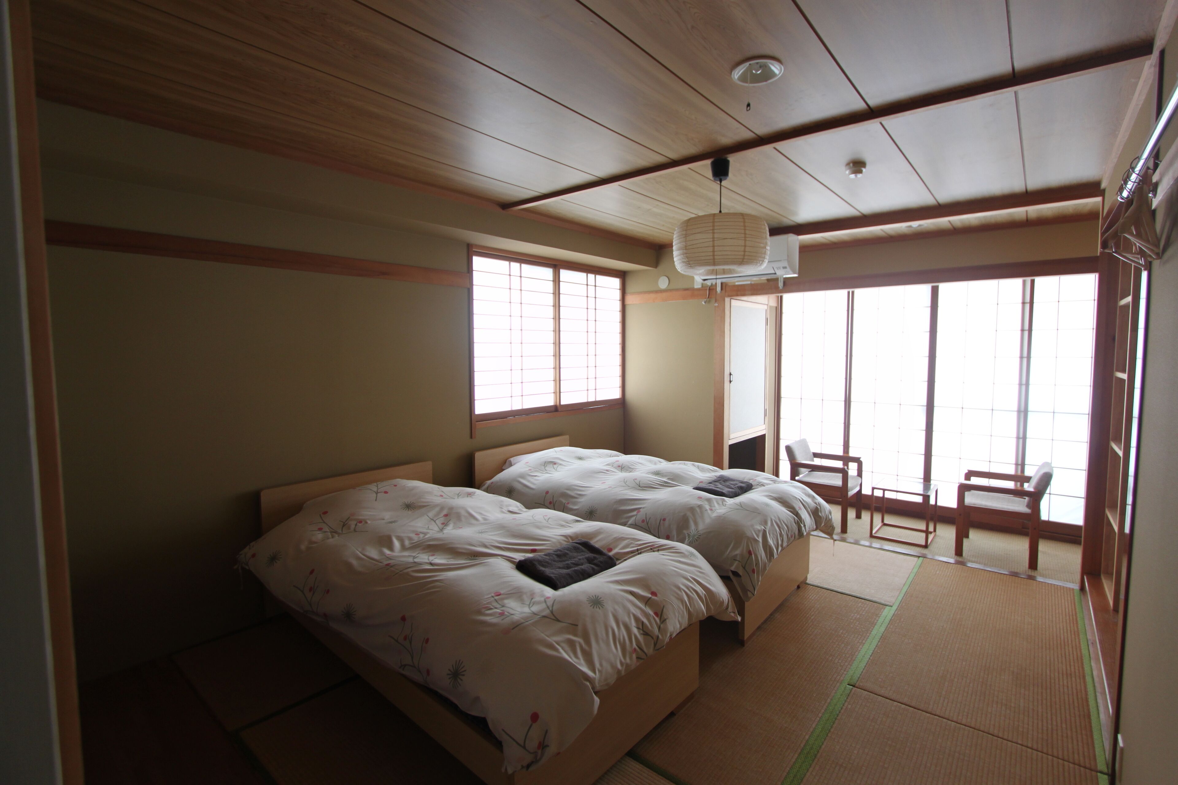 A traditional Japanese-style room with tatami flooring, two beds with white bedding, a wooden ceiling, and large windows providing natural light.
