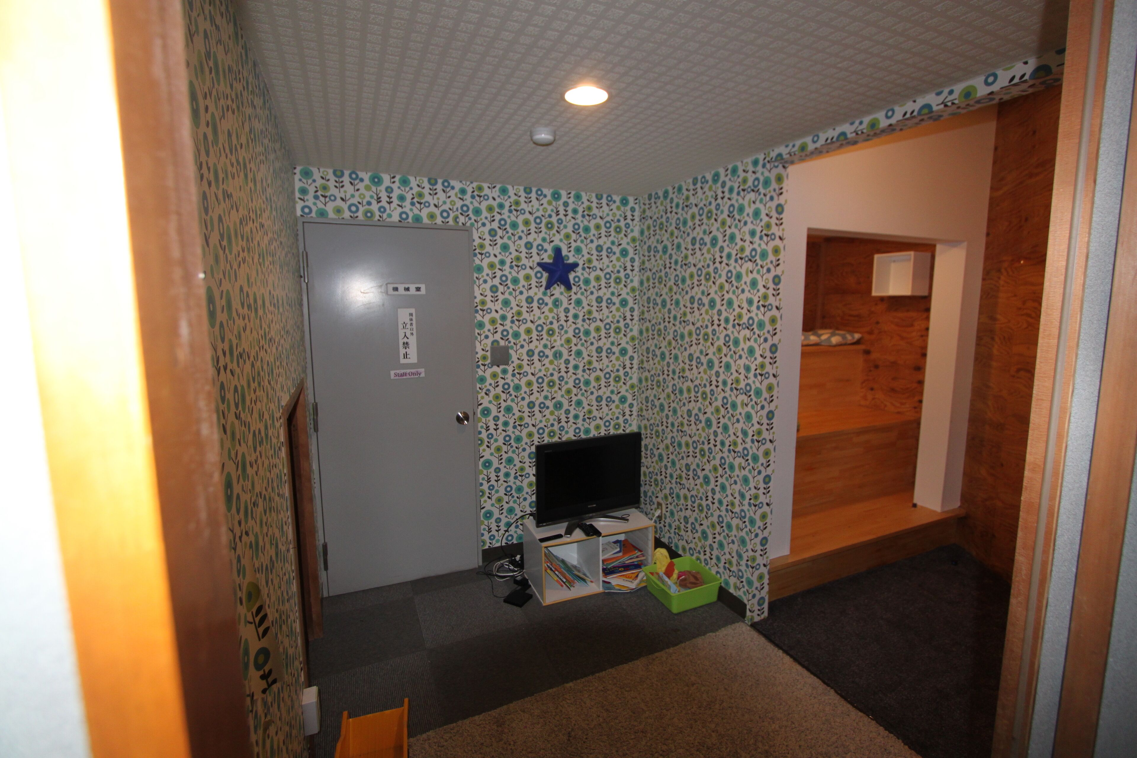 A room with patterned wallpaper, a television on a stand, a door, and a small wooden shelf.
