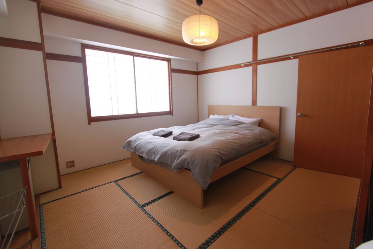 A room with tatami flooring, a wooden bed with a headboard, a window with blinds, and a wooden wardrobe.