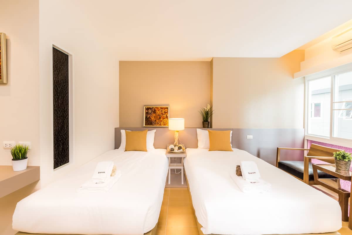 A hotel room with two single beds, a nightstand with a lamp, a small table with a plant, and a chair.
