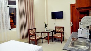 Habitación de hotel con cama, una pequeña mesa de comedor con dos sillas, televisión montada en la pared, zona de cocina con lavamanos y cafetera, y ventana con cortinas.