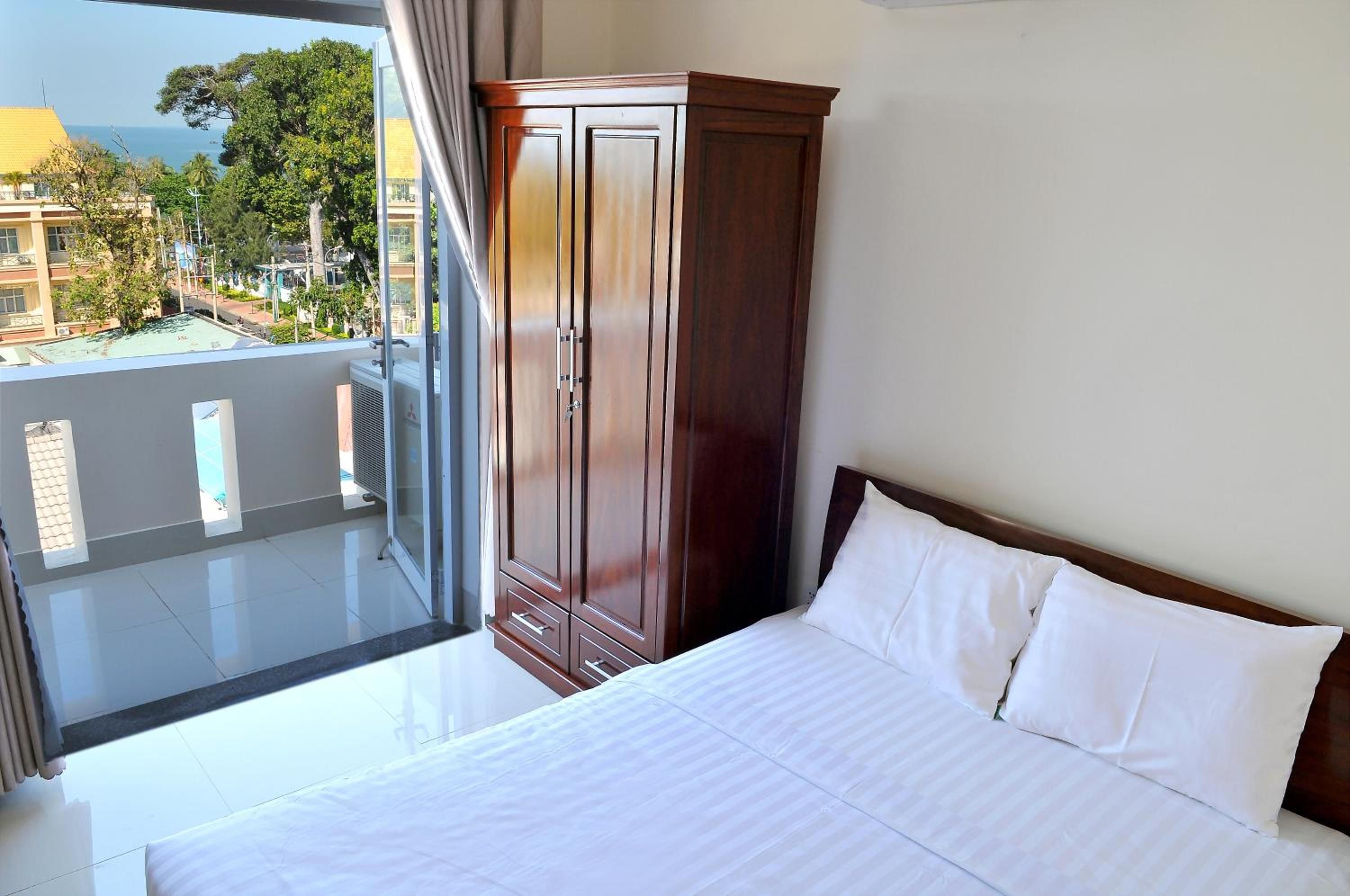 Une chambre d’hôtel avec un lit, une armoire en bois et un balcon donnant sur des immeubles et des arbres.