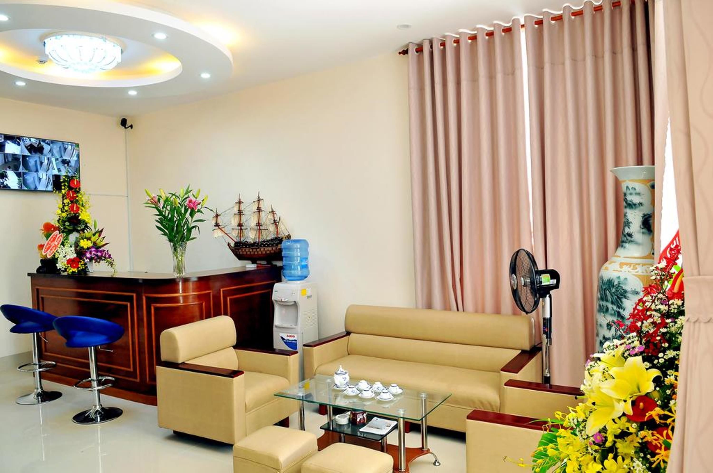 A modern living room with a beige sofa, a glass coffee table, and blue bar stools. There is a wooden reception desk with a ship model and a water dispenser.
