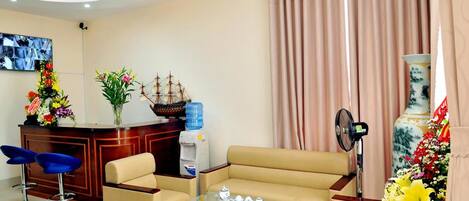 Una sala de estar moderna con un sofá color beige, una mesa de centro de vidrio y taburetes azules. Hay un mostrador de recepción de madera con un modelo de barco y un dispensador de agua.
