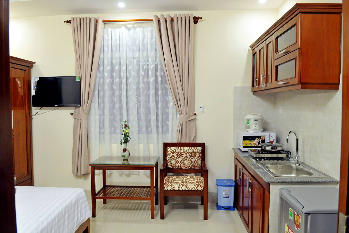 Ein kleines Hotelzimmer mit Bett, einem Tisch mit Stuhl, einer Küchenzeile mit Spüle und einem an der Wand befestigten Fernseher.