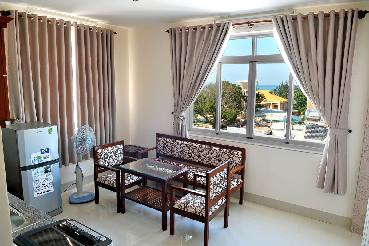 Ein Zimmer mit Esstisch, Stühlen, einem Kühlschrank, einem Ventilator und einem Fenster mit Vorhängen.