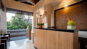 Un espace d’accueil doté d’un comptoir de réception en bois, d’une grande fenêtre avec des stores et d’un mur décoré d’un motif texturé et agrémenté de plantes en pot.