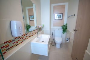 Un baño moderno con un lavamanos blanco, un inodoro y una ducha. Hay un dispensador de jabón en la pared y un estante pequeño con artículos de aseo.