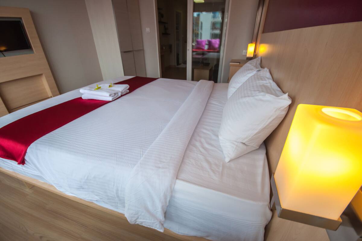 Una cama bien hecha con sábanas blancas y una manta roja, una lámpara de noche y un cabecero de madera.