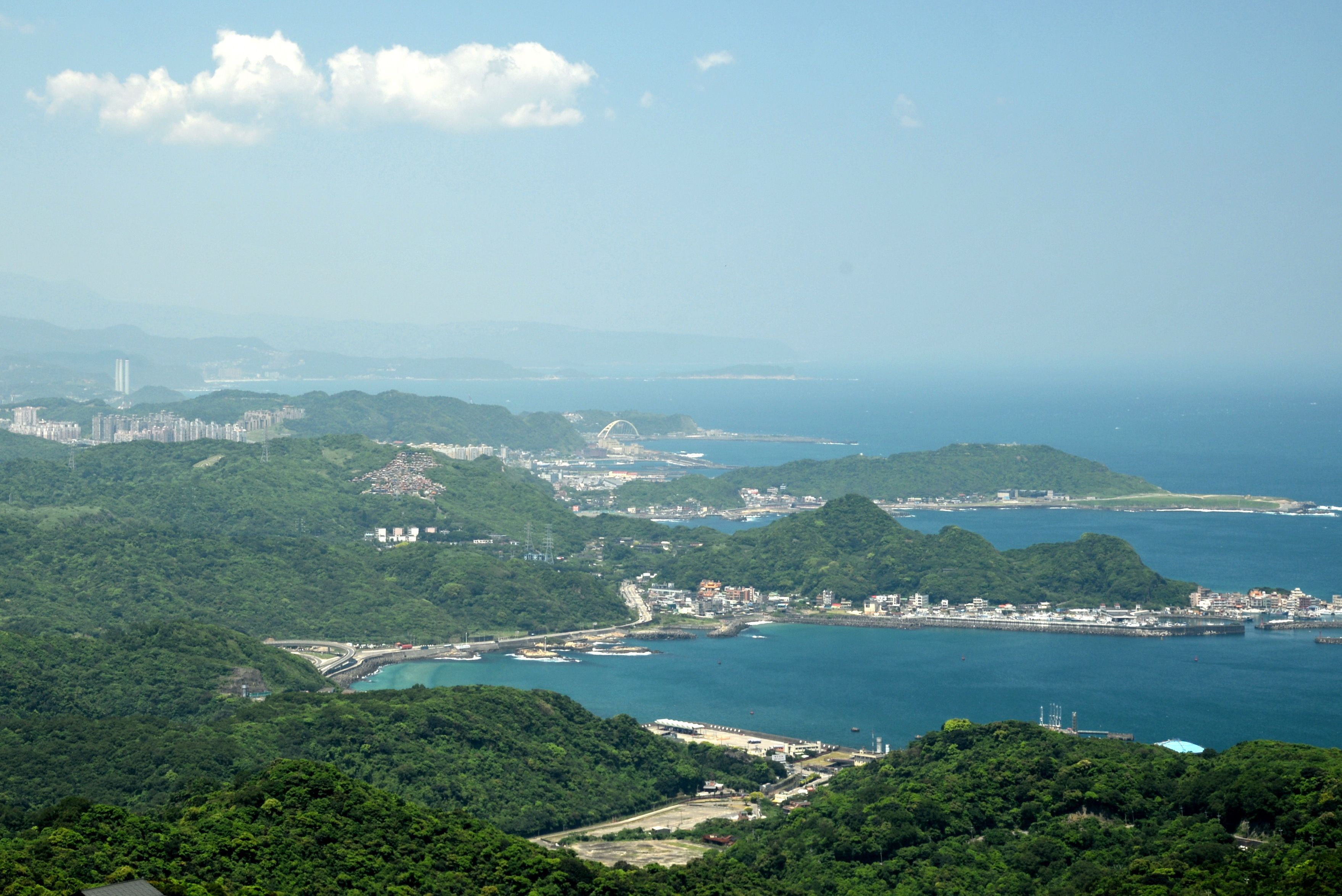 A coastal cityscape with dense greenery, a marina, and a distant bridge.