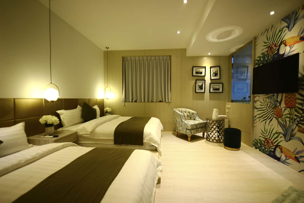 A hotel room with two beds, a chair, a side table with a lamp, and a wall with framed pictures.