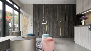 A modern lobby with a round metallic table, a pink ottoman, and a blue stool. A large dark wooden door with vertical slits is visible in the background. - Happy House B&B, Juifan (New Taipei City)