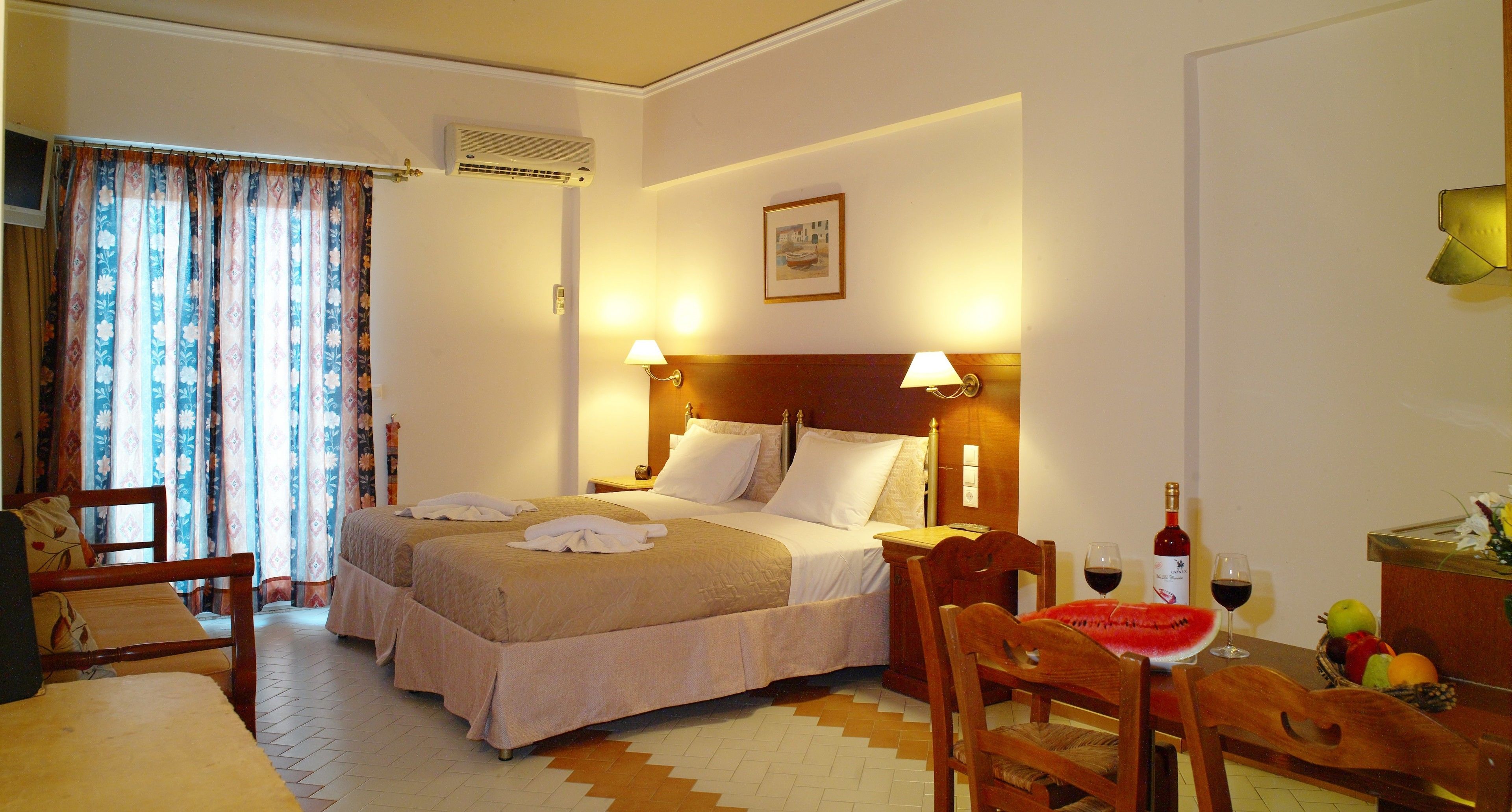 A hotel room with a double bed, a wooden headboard, two bedside lamps, a wooden nightstand, a chair, a table with a fruit bowl and wine bottle, and a window with curtains.