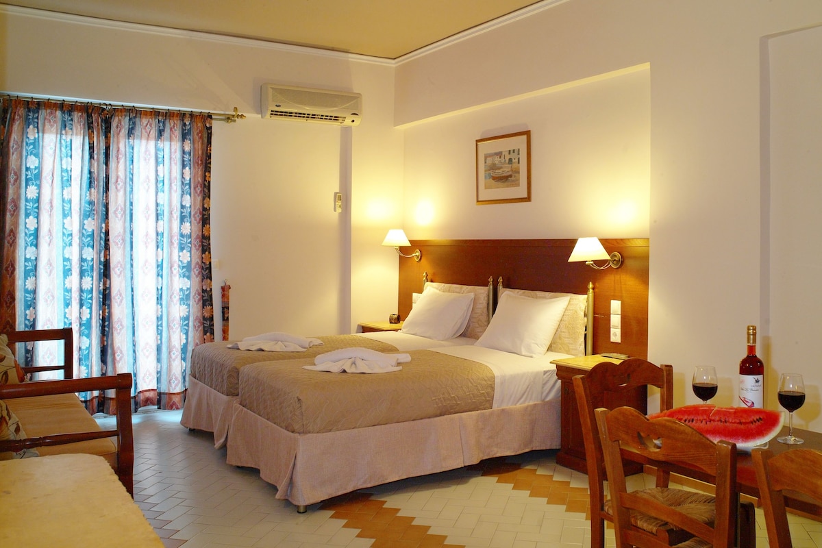 Hotelkamer met een tweepersoonsbed, een houten hoofdbord, twee bedlampjes, een nachtkastje van hout, een stoel, een tafel met een schaal fruit en een fles wijn, en een raam met gordijnen.