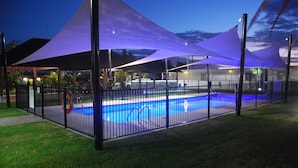 A swimming pool with a fence and shaded areas, surrounded by grass and palm trees. - Cherratta Lodge (Karratha Industrial Estate)