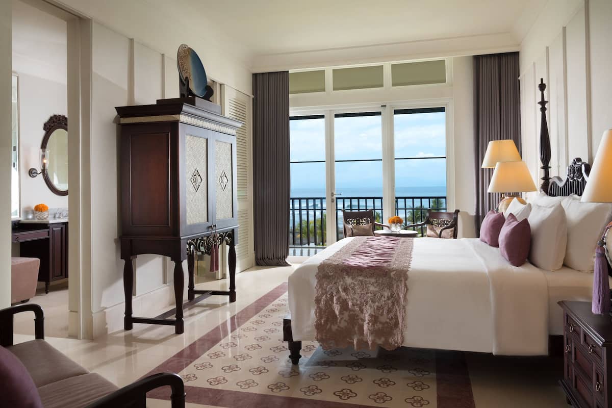 A hotel room with a large bed, a dresser, a desk with a mirror, and a view of the ocean through large windows.