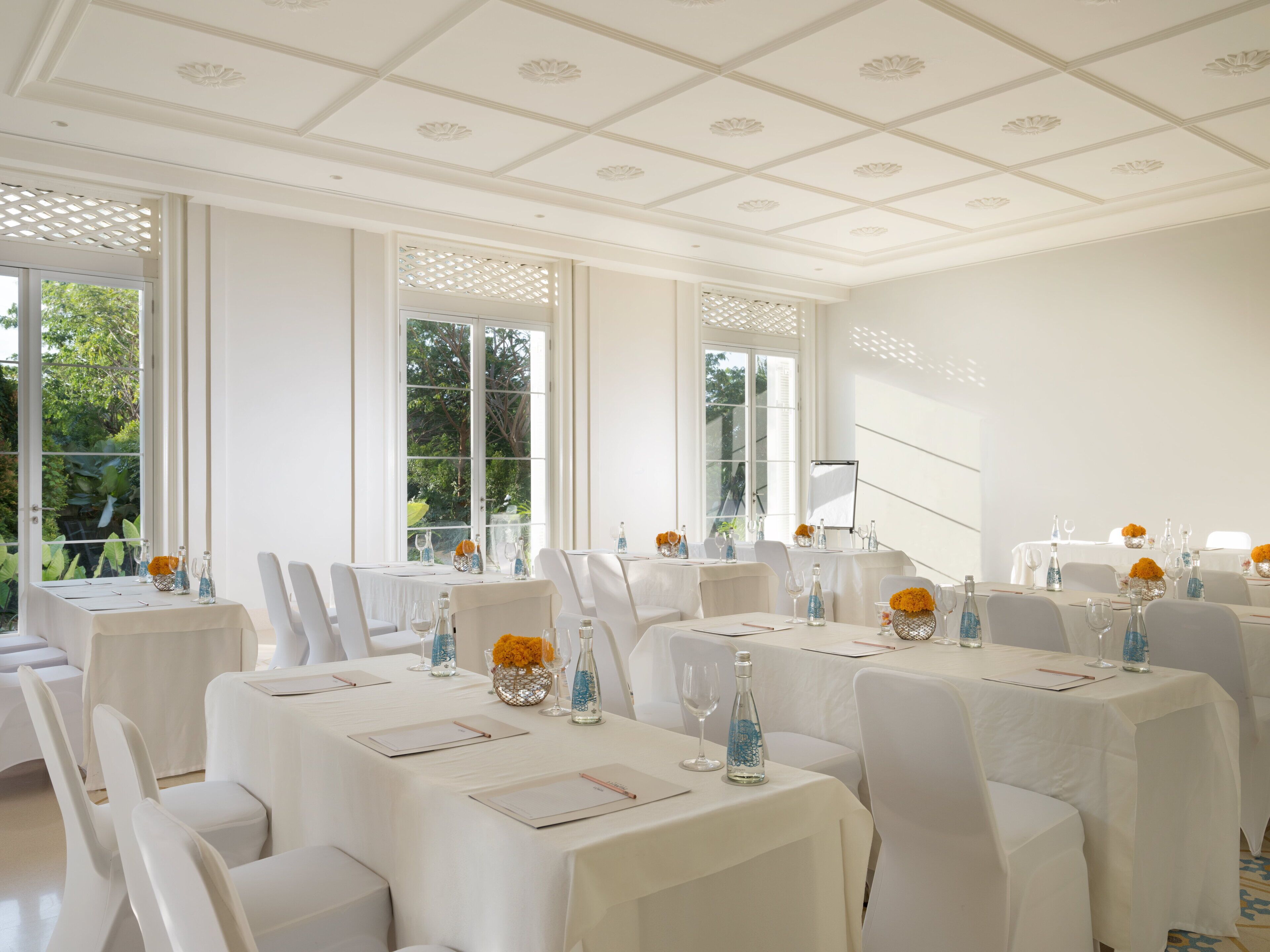 Ein geräumiger Raum mit mehreren Esstischen, großen Fenstern mit Blick auf Grünflächen und einer weißen Decke mit dekorativen Paneelen.