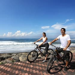 Cyclists ride along the oceanfront walkway, surrounded by large rocks and a brick pattern.