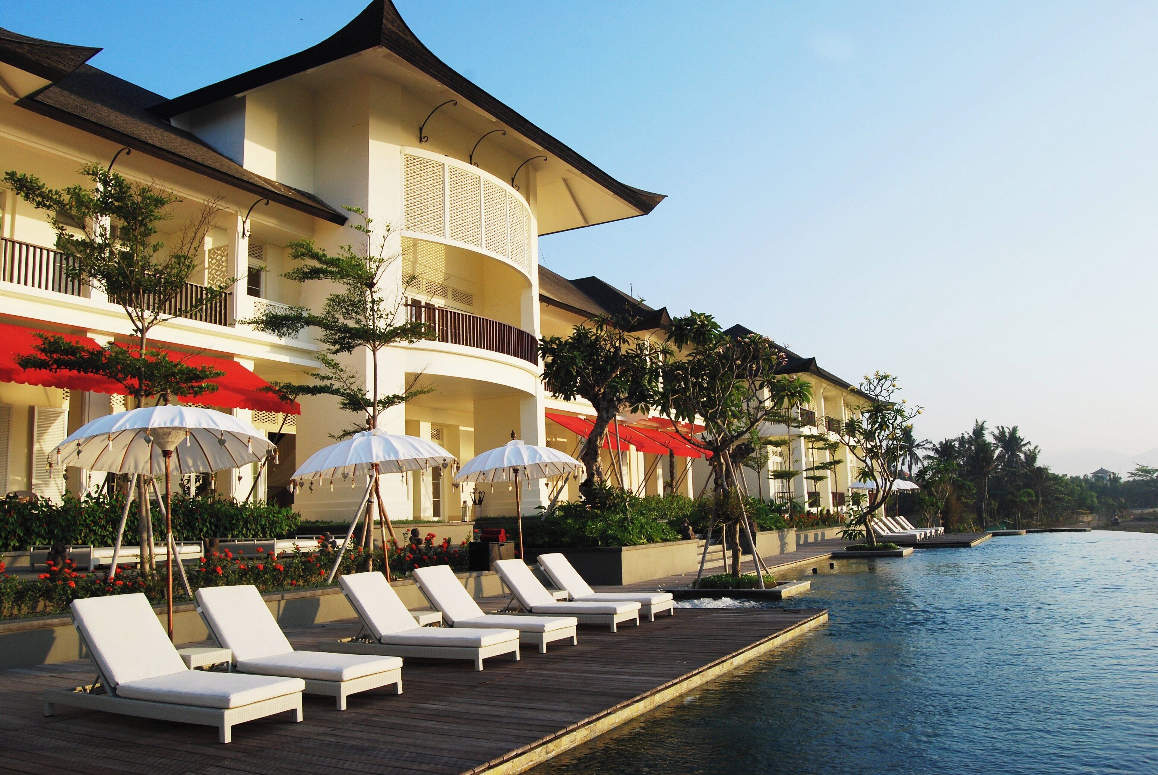 A resort with a poolside lounge area, white sun loungers, and red umbrellas.