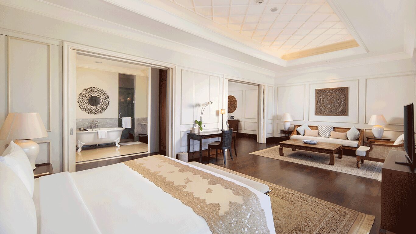 Une chambre d’hôtel spacieuse, dotée d’un grand lit, d’un coin salon avec un canapé et une table basse, d’un bureau avec une chaise, et d’une salle de bain visible à travers une porte ouverte.