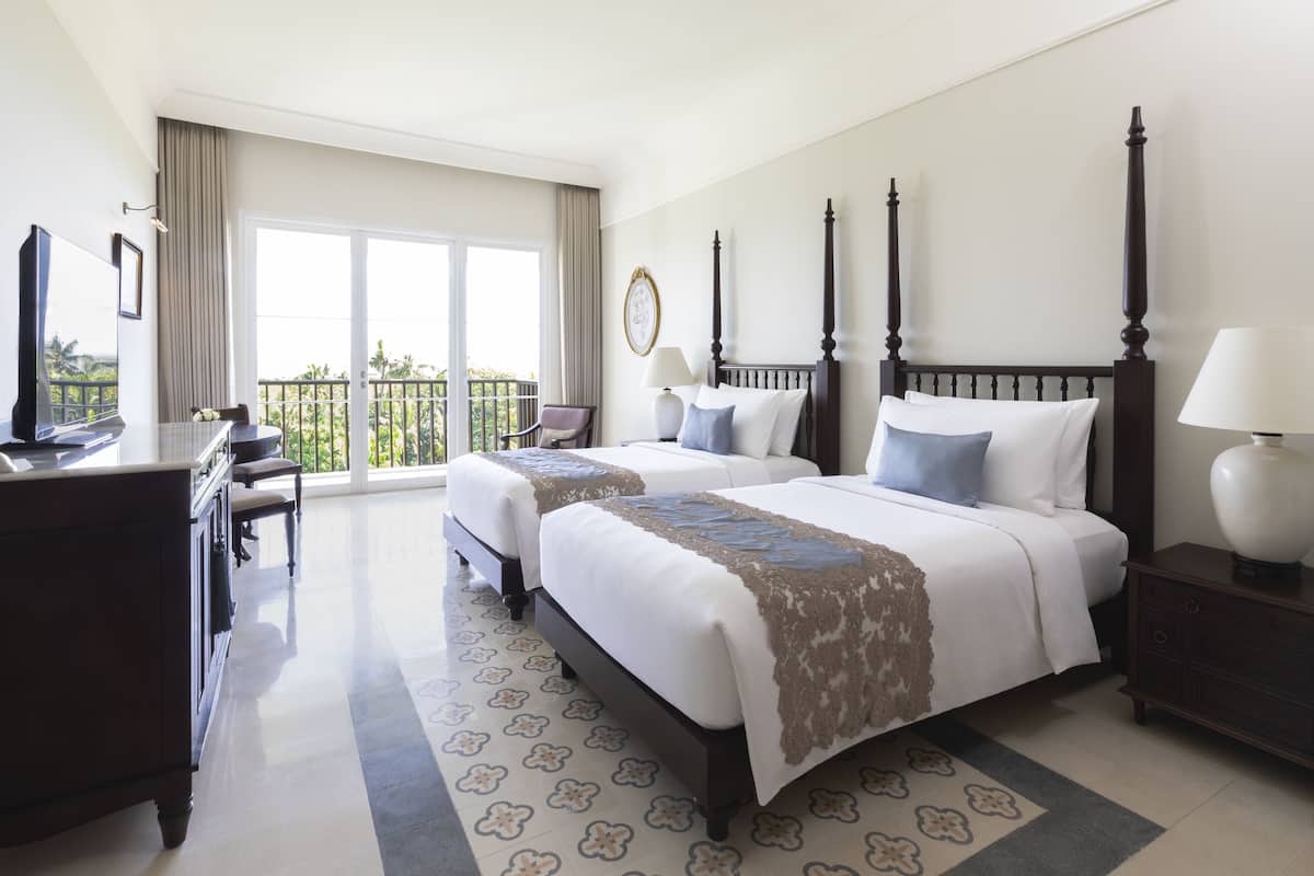 A hotel room with two beds, a desk, a chair, a TV, and a balcony with a view.