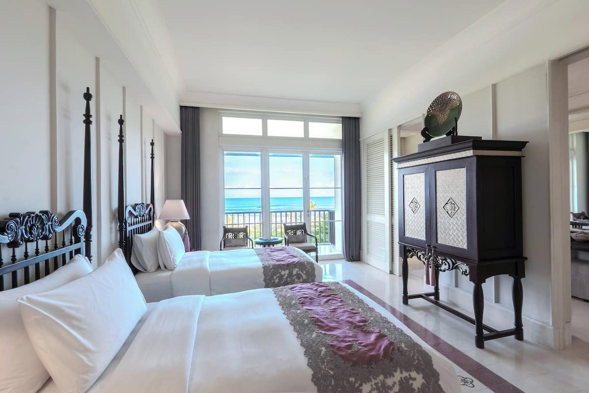 A hotel room with two beds, a view of the sea, a large window, and a cabinet with decorative panels.