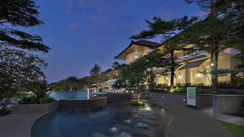 Uma piscina com design curvo, rodeada por espreguiçadeiras e um edifício com estilo arquitetônico tradicional.