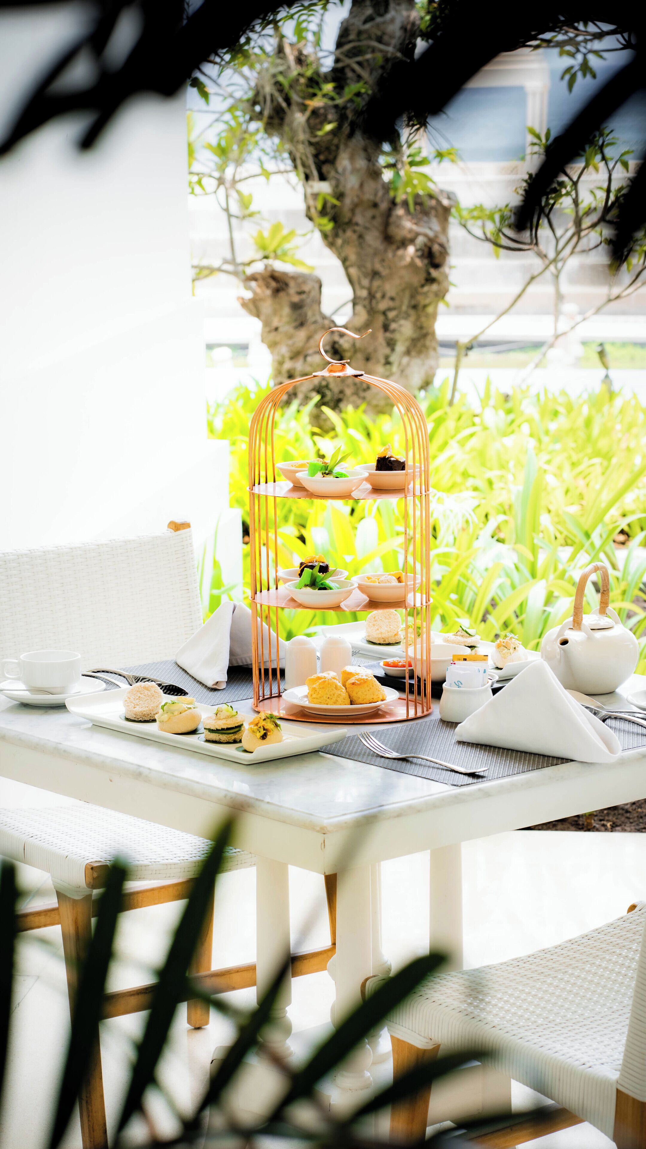 A three-tiered dessert stand with various pastries and a teapot on a table set outdoors with a view of greenery and a tree.