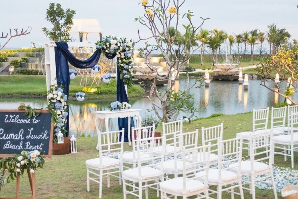 An outdoor wedding setup with white chairs arranged in rows, a sign welcoming guests to Rumah Luwih Beach Resort, and a decorative arch with flowers.