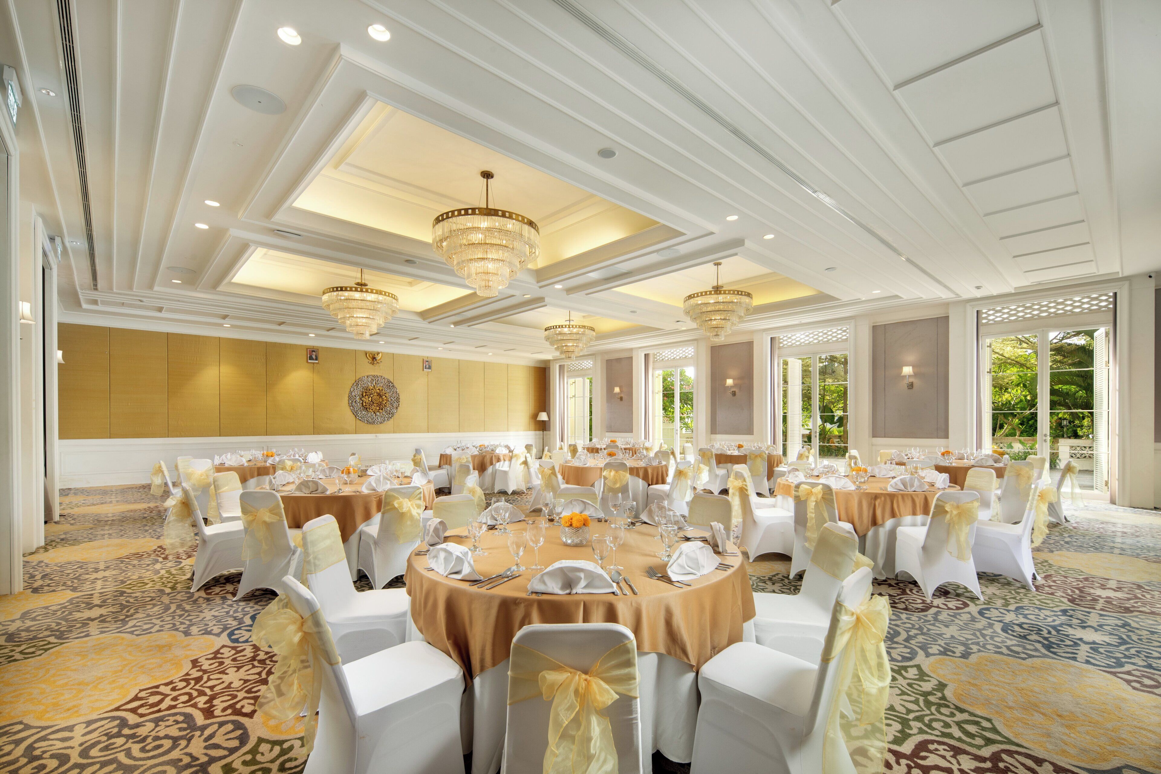 A banquet hall with round tables, white chairs, chandeliers, and a patterned carpet.