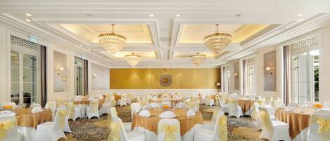 A banquet hall with round tables, white chairs with gold sashes, chandeliers, and a patterned carpet.