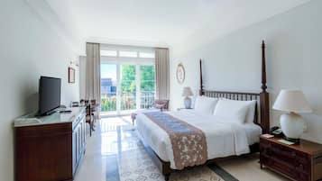 A hotel room with a large bed, a TV, a desk, and a balcony with a view of greenery.
