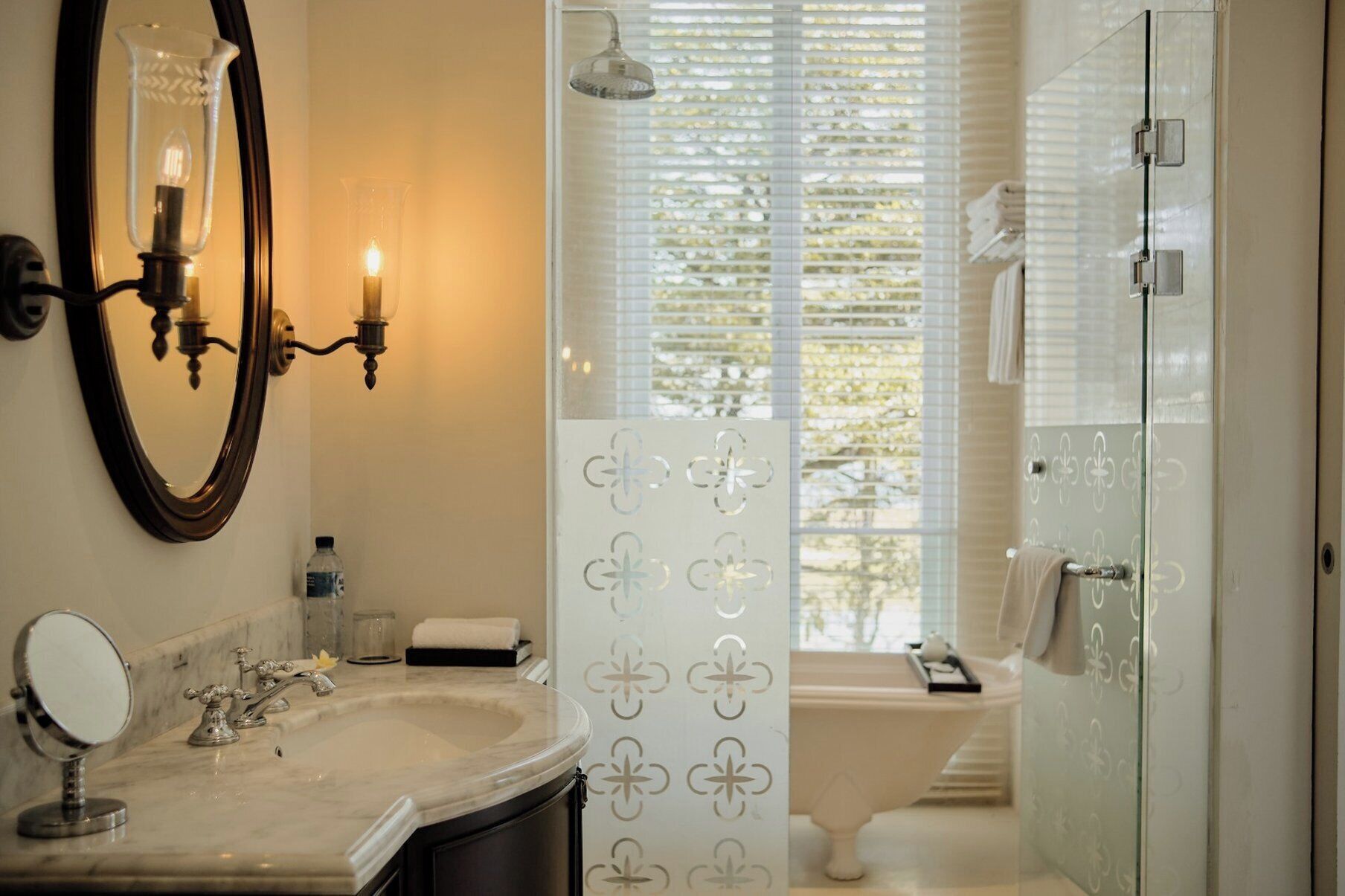 A bathroom with a marble sink, a round mirror, a wall-mounted light fixture, and a shower with a glass door.