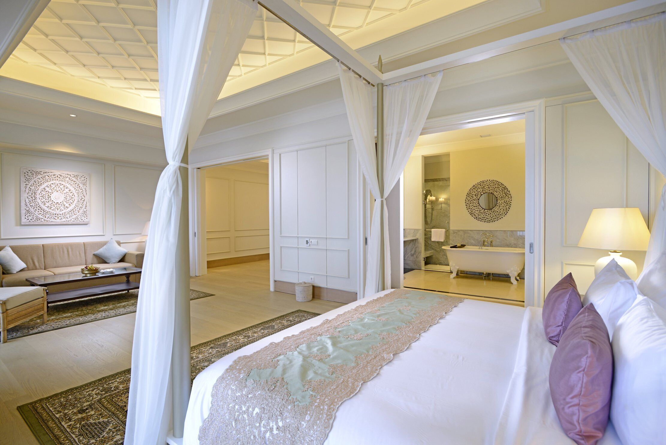 A bedroom with a four-poster bed, a sitting area with a sofa and coffee table, and a bathroom with a bathtub and a mirror.