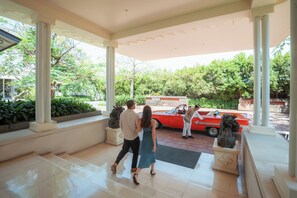 A couple walking towards a red car parked outside a building with columns and a tiled floor. - Rumah Luwih Bali (Gianyar)