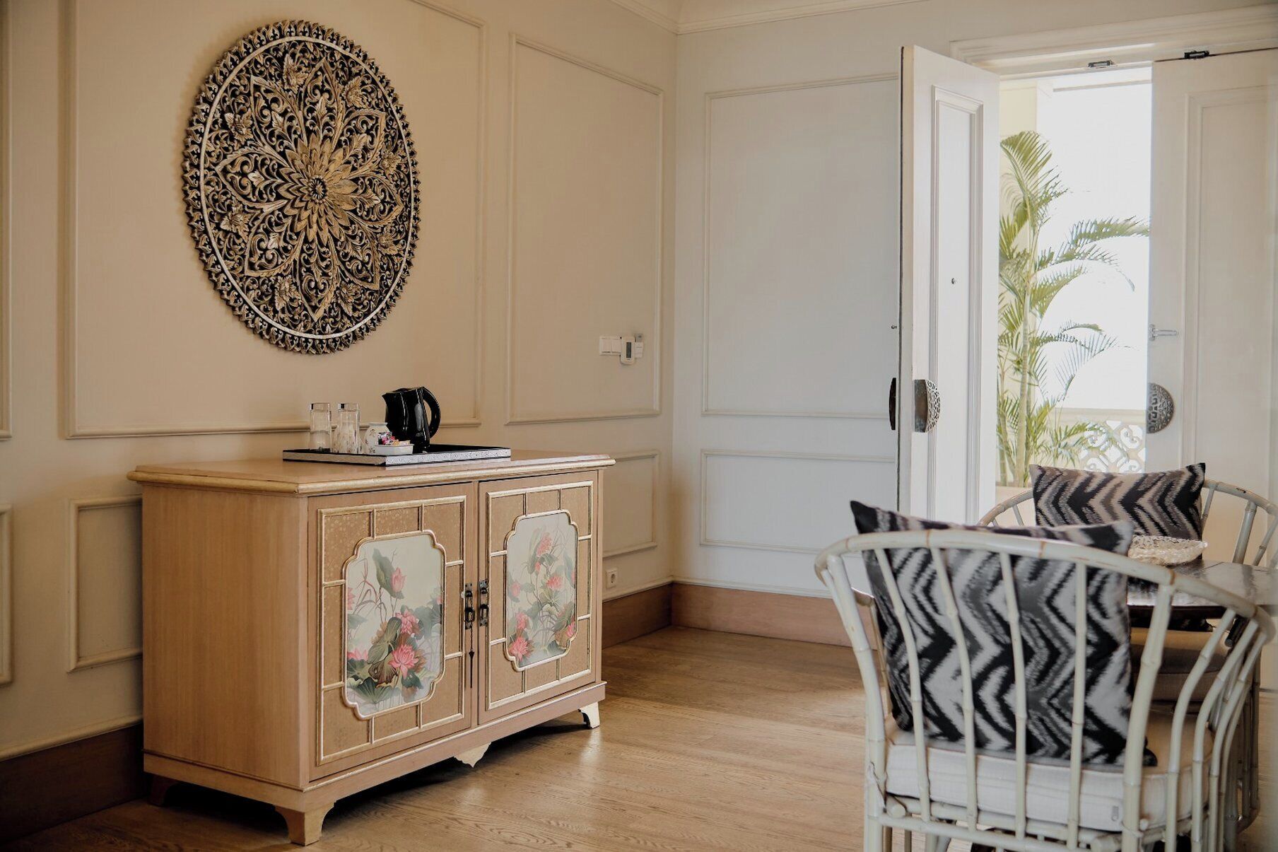 A room with a wooden cabinet with floral designs, a white chair with patterned cushions, and a decorative wall piece.