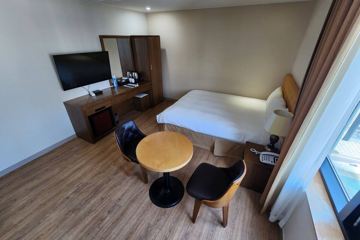 A hotel room with a bed, a desk with a TV, a chair, a small table, and a window with curtains.