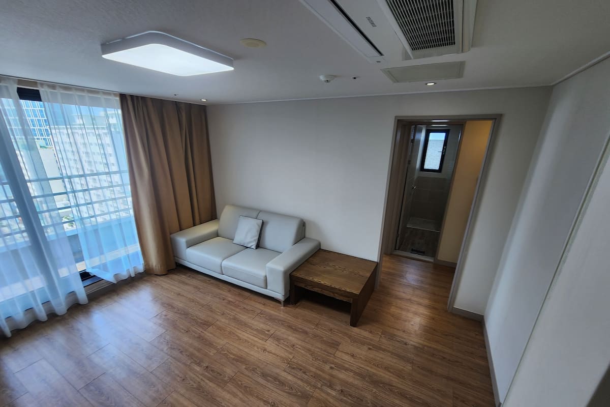 A modern living room with a light-colored sofa, a wooden coffee table, and a view through large windows with sheer curtains.