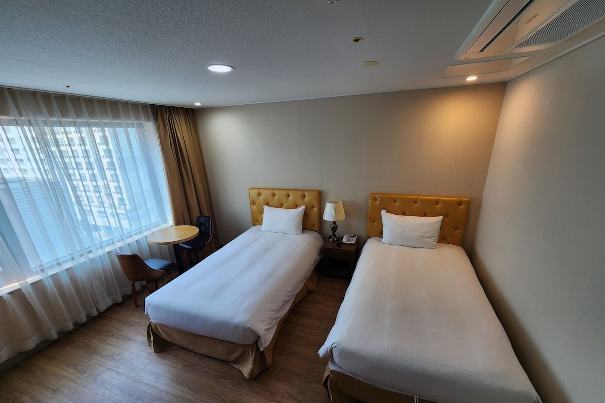 A hotel room with two single beds, a small round table, a chair, and a window with curtains.