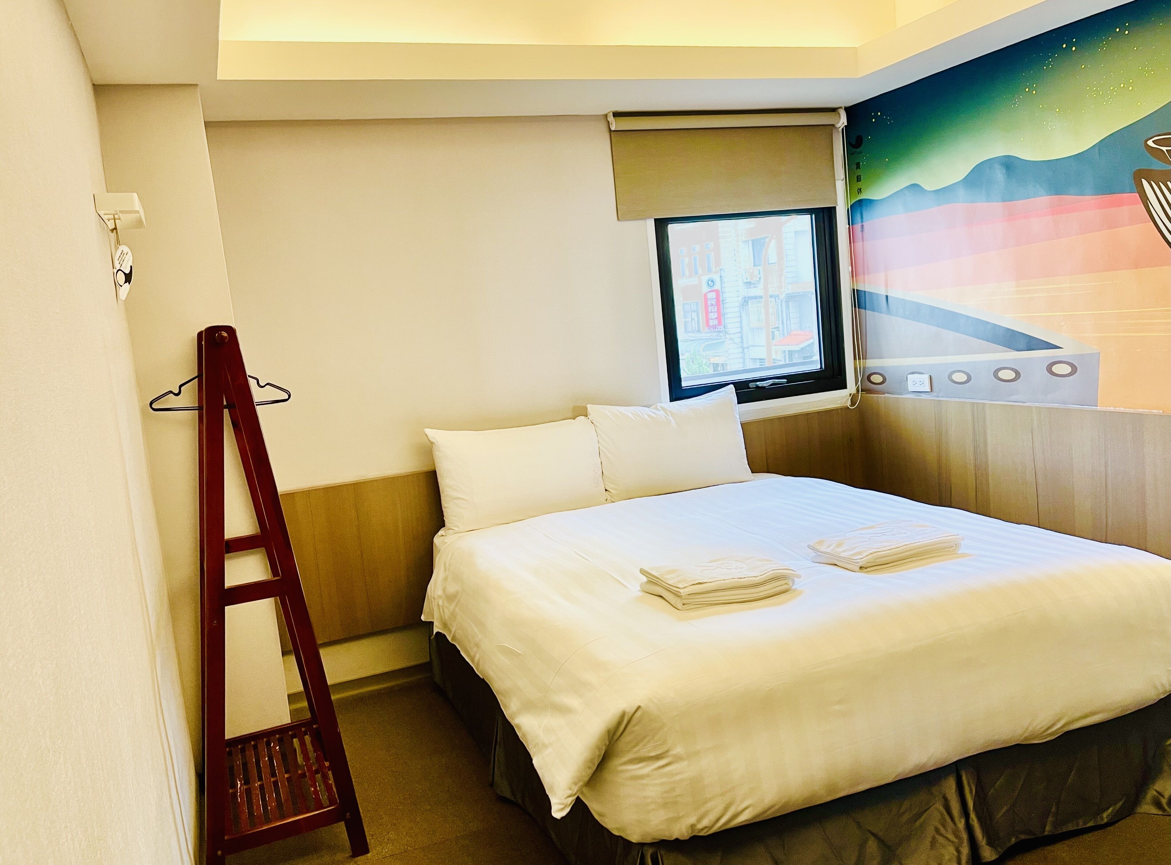 A hotel room with a bed, a wooden headboard, a window with a view of buildings, a wall-mounted TV, and a wall decoration.