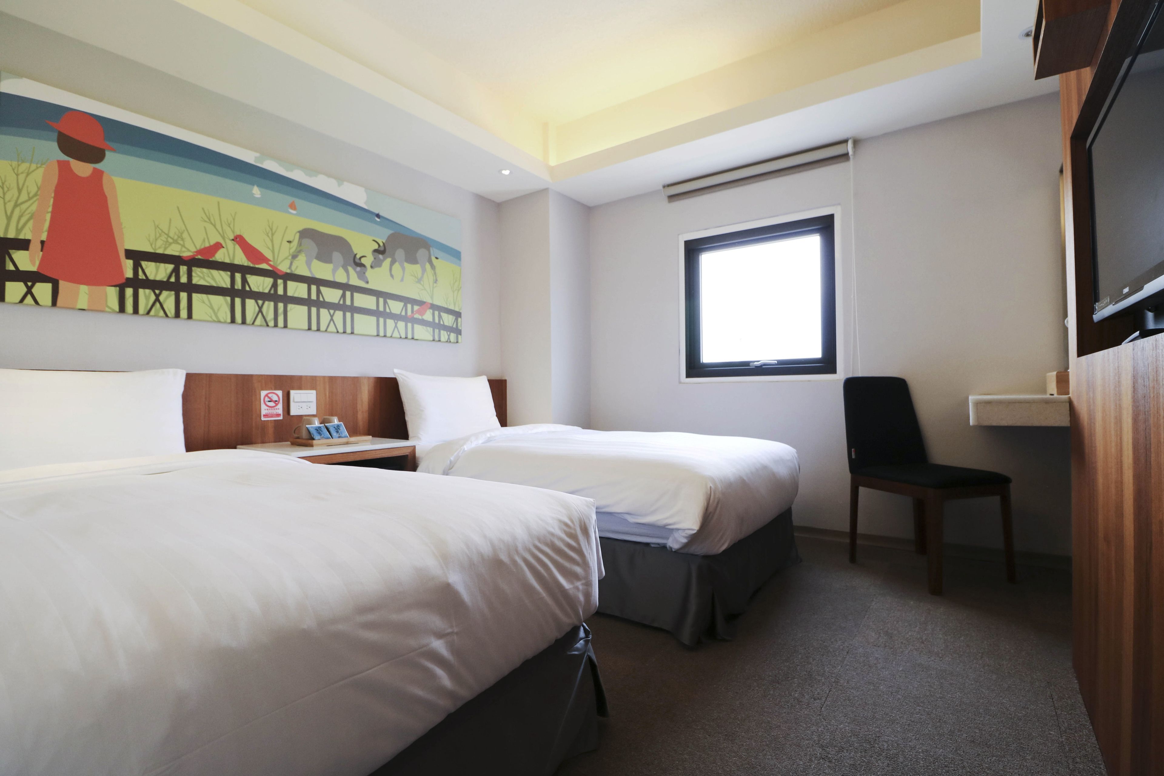 A hotel room with two beds, a desk, a chair, a TV, a window, and a mural on the wall.