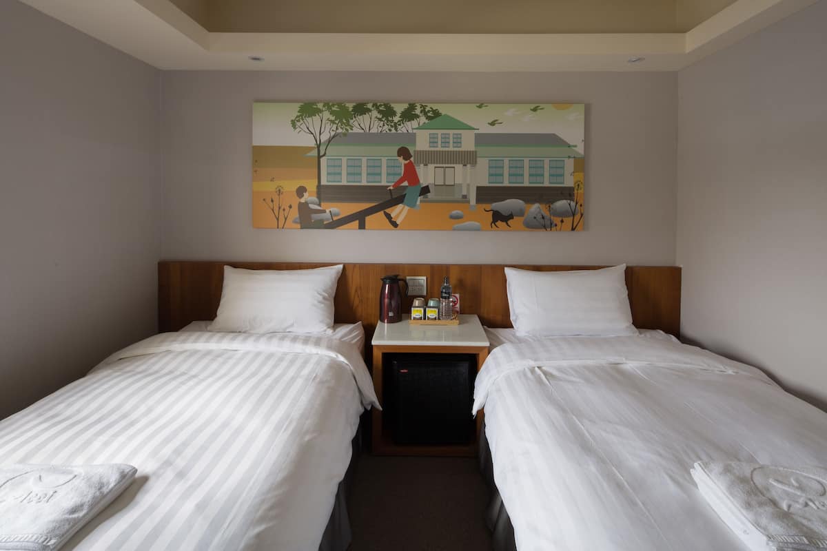 A hotel room with two beds, a nightstand, a painting, and a small refrigerator.