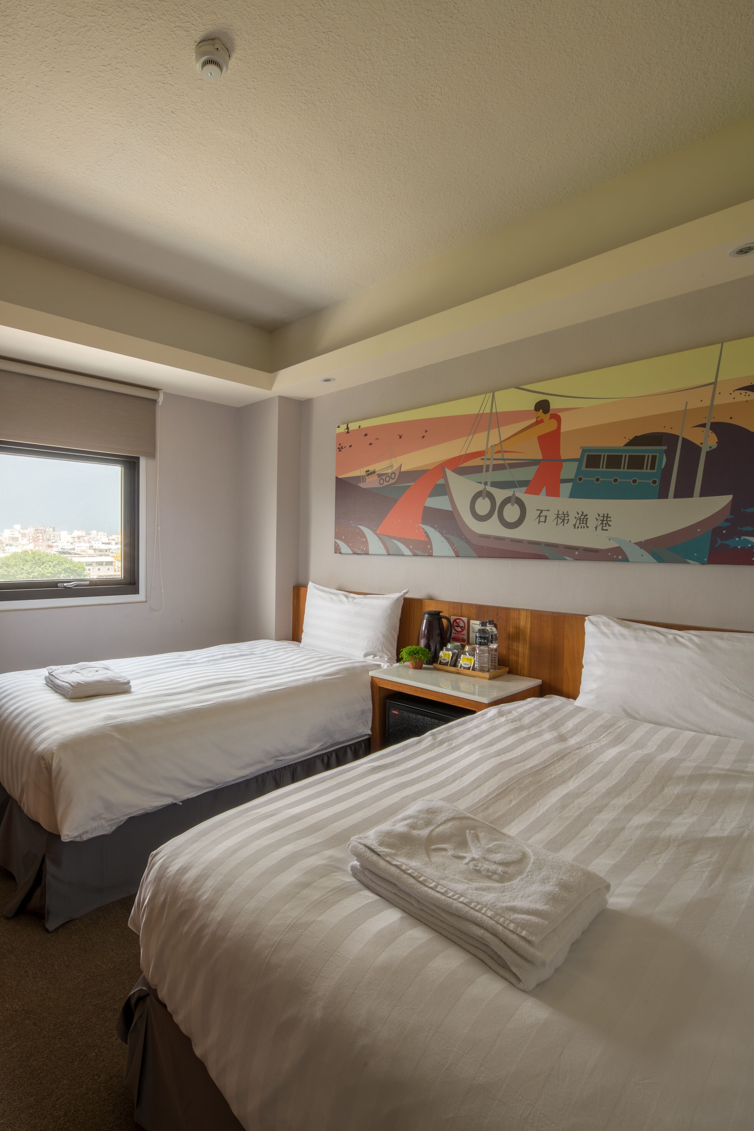 A hotel room with two beds, a window, a nightstand with a lamp, and a wall mural.