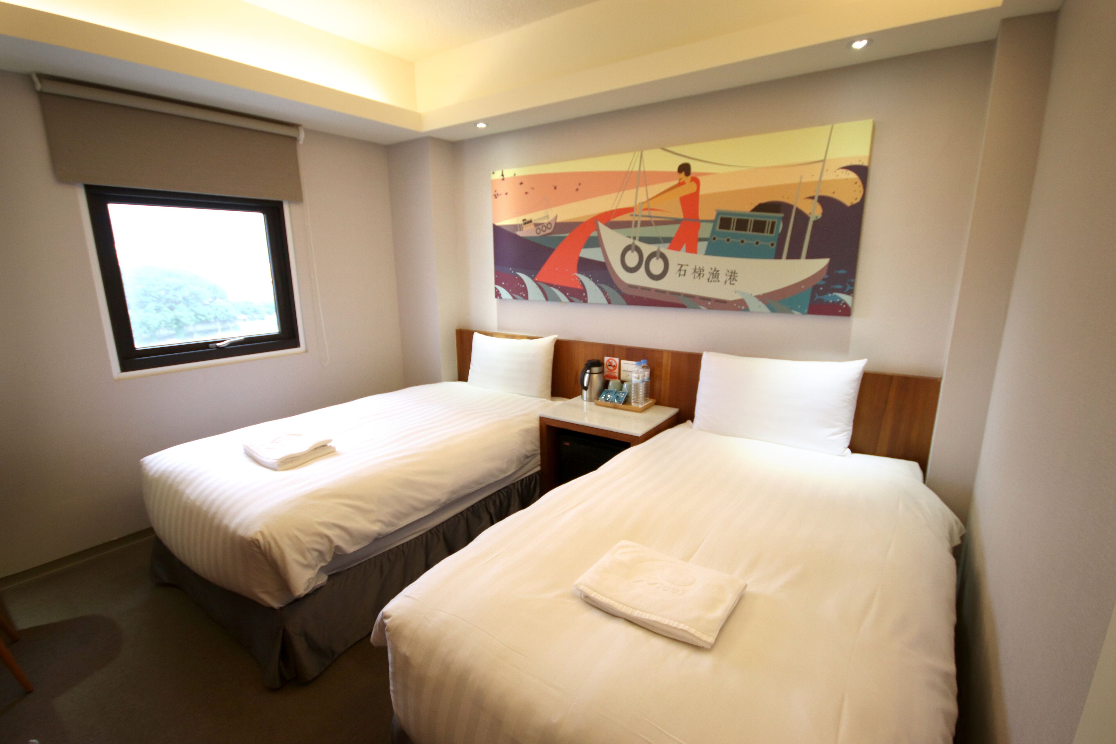 A hotel room with two beds, a window, and a painting on the wall.