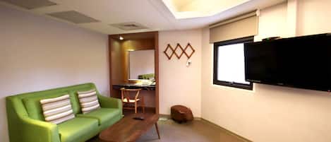Una habitación con un sofá verde, una mesa de centro de madera, un televisor en la pared y un escritorio pequeño con una silla.